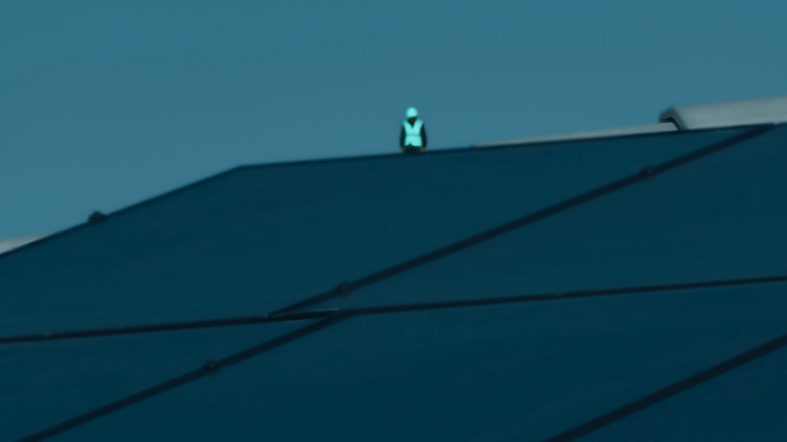 Close-up of dark solar panels on a rooftop with a small figure wearing a bright cyan reflective vest and hard hat visible in the background against a clear blue sky.